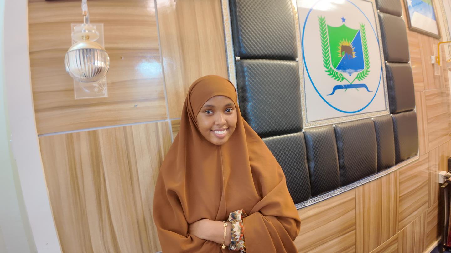 Smiling woman in brown hijab stands in a wood-paneled lobby with a crest on the wall.