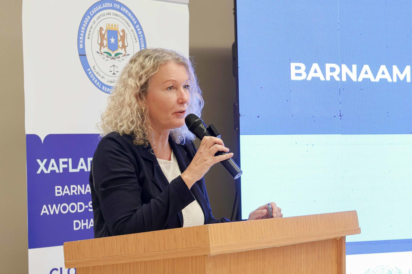Woman speaking at a wooden podium with microphone during a conference, blue banners and screen behind.
