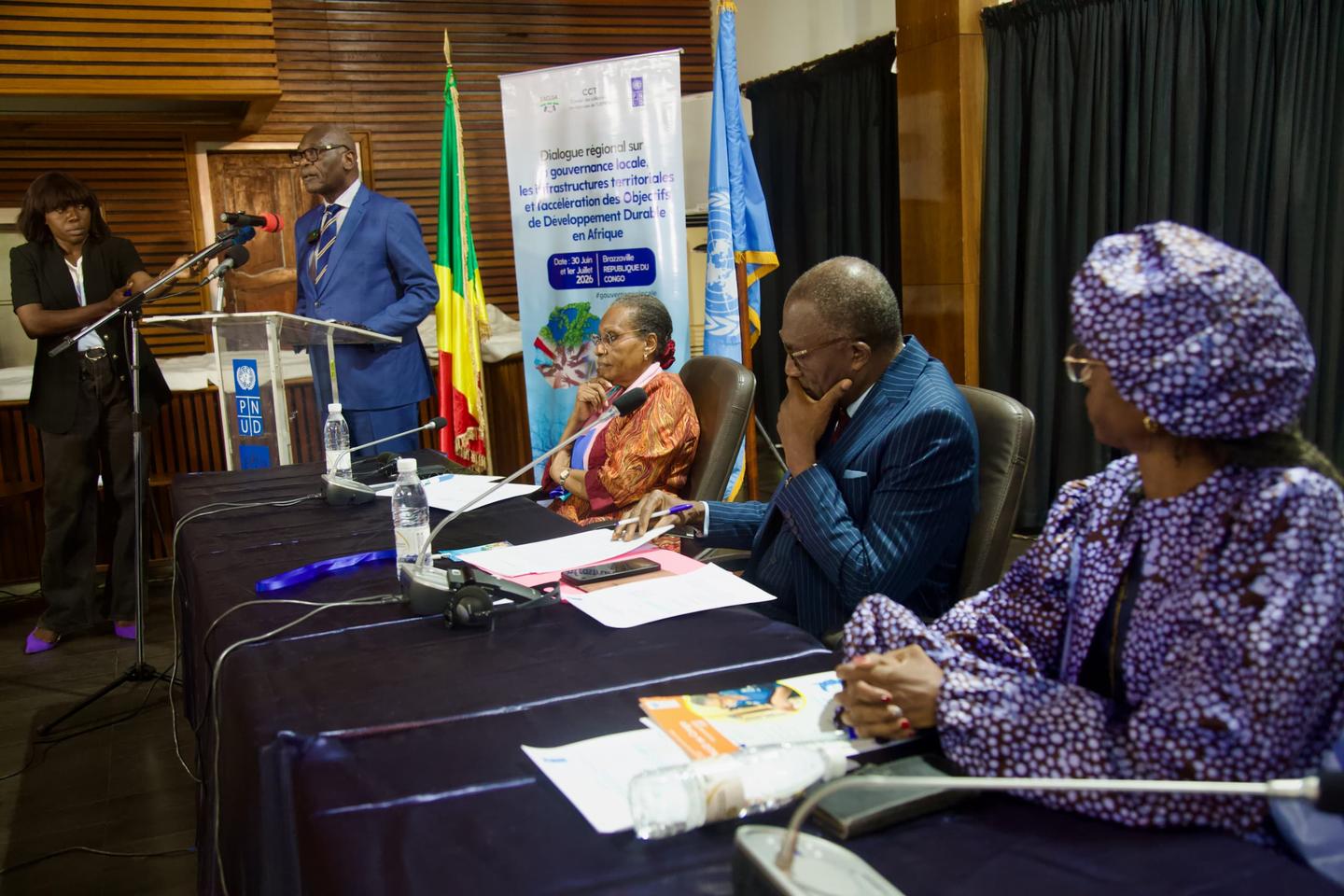 Panelists sit at a long table on a stage; a man in a blue blazer stands at a podium beside a banner.