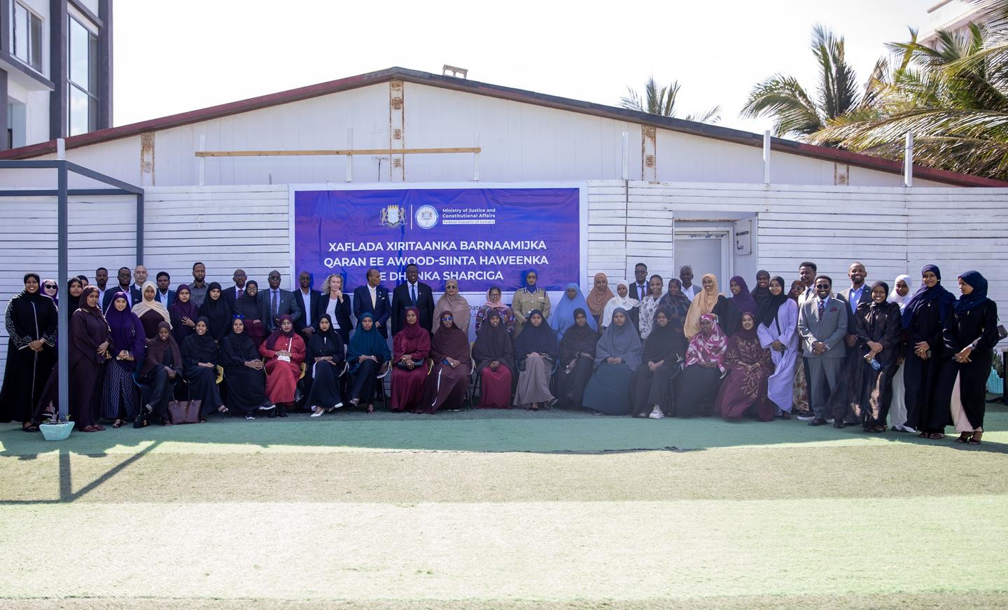 Group photo of students and staff outside a light-blue building with a purple banner.