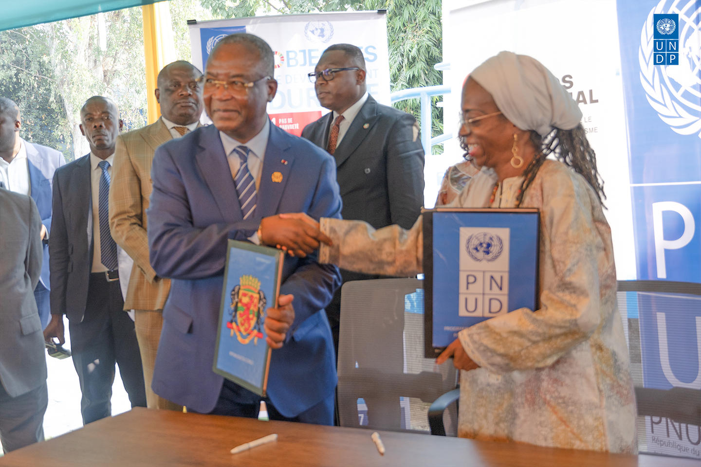 Man in suit and woman in hijab shake hands as a blue folder is exchanged at a formal signing event.