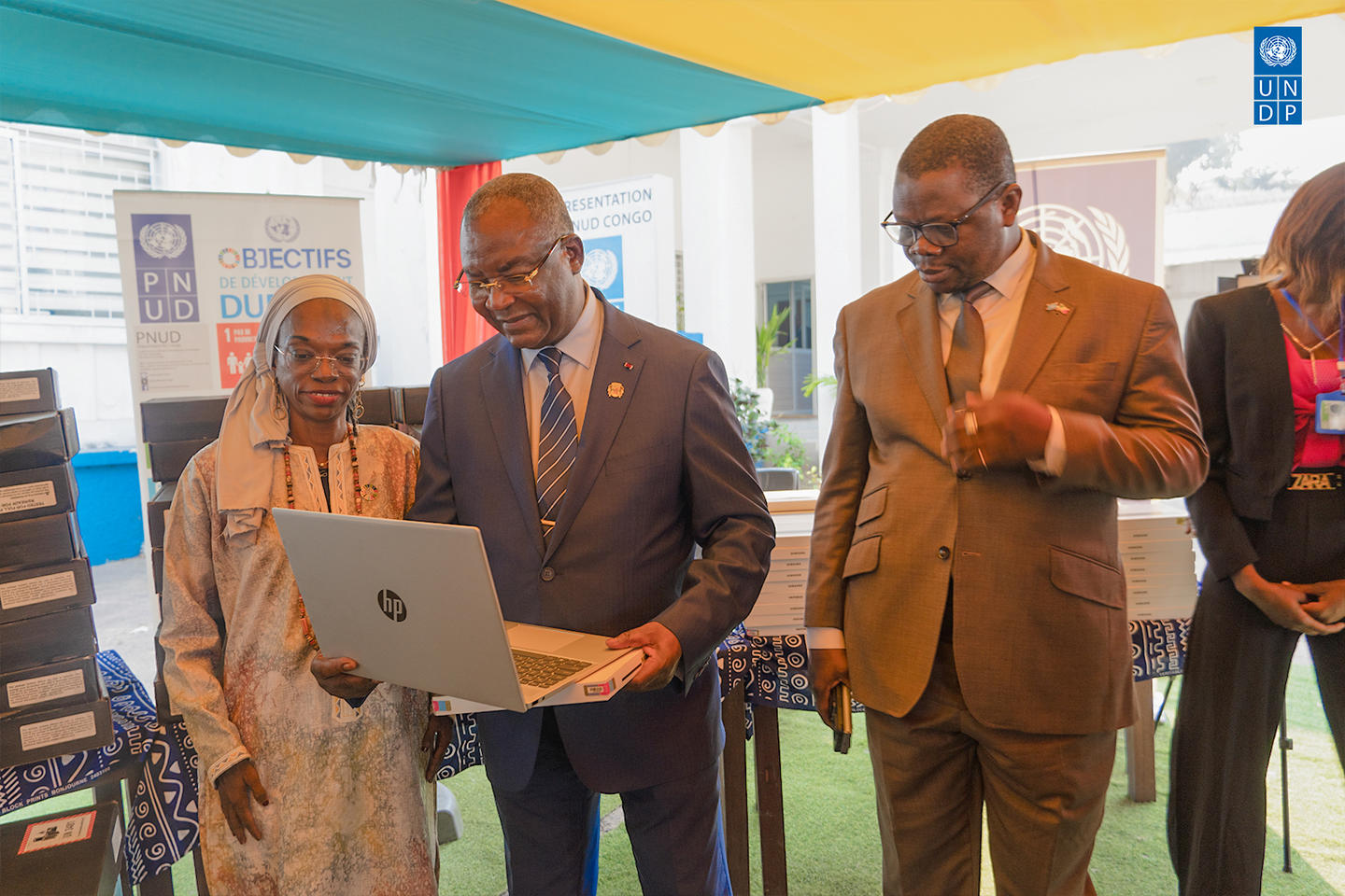 Group of four adults under a yellow-blue canopy; man in blue suit presents a laptop.