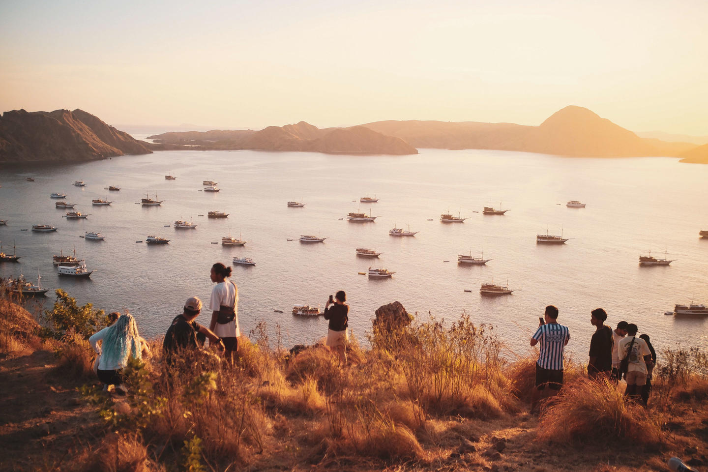 Sunset over a bay with many boats; people stand along a grassy cliff in the foreground.
