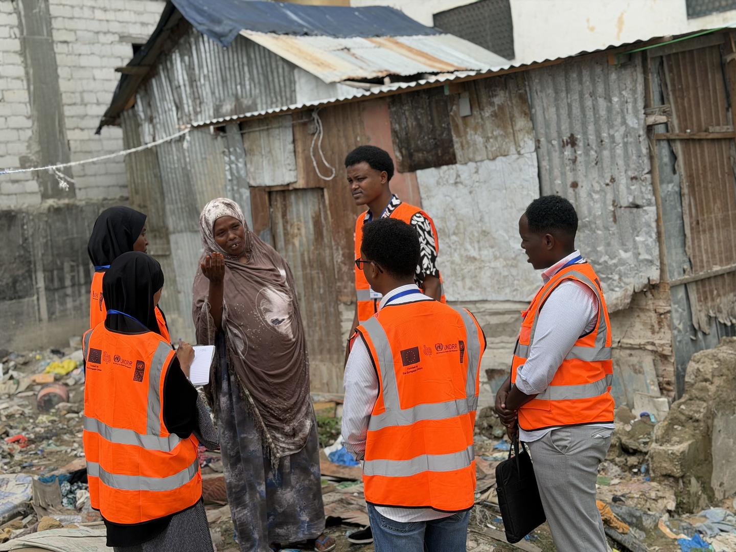 Group of people in orange safety vests talking near rundown wooden shacks