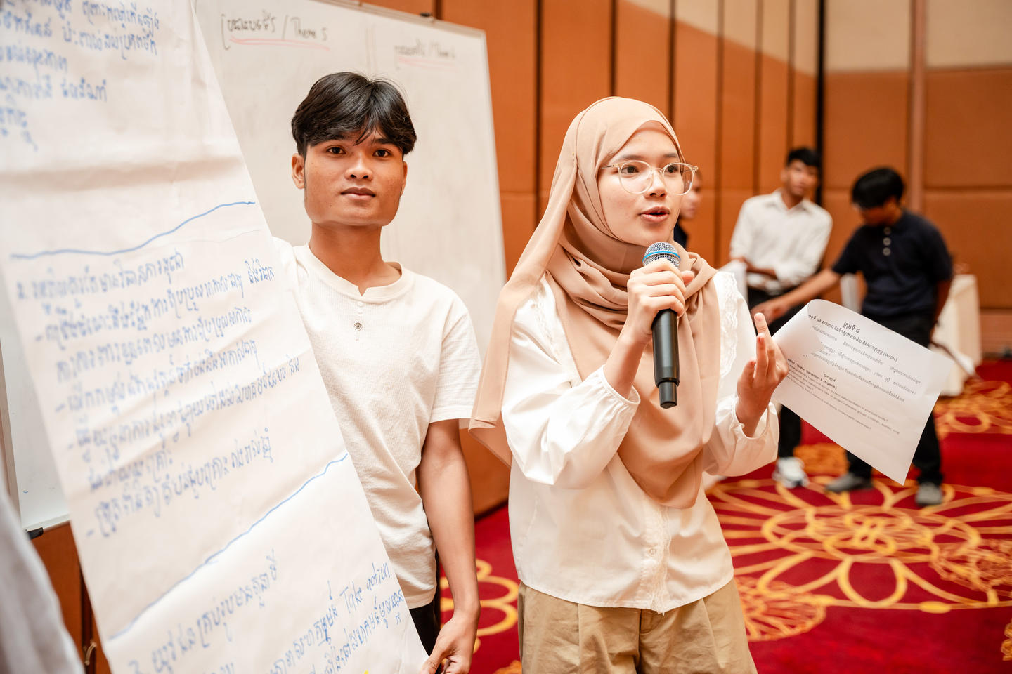 Photograph: girl in hijab speaks into microphone beside boy at a workshop, whiteboard behind.