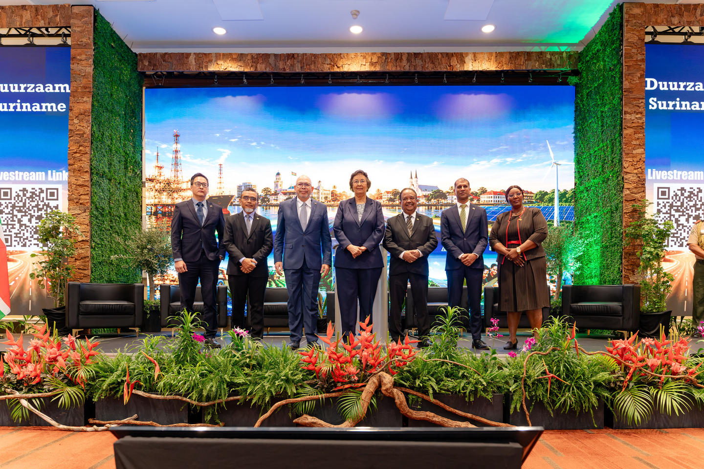 Group of people in suits standing on a stage with a cityscape backdrop and greenery.
