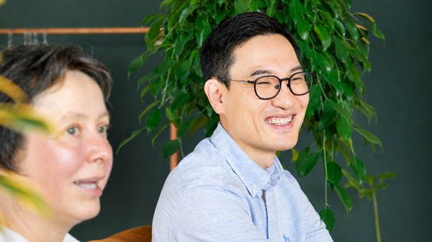 Two coworkers in an office; woman on the left, man with glasses smiling, plant behind.