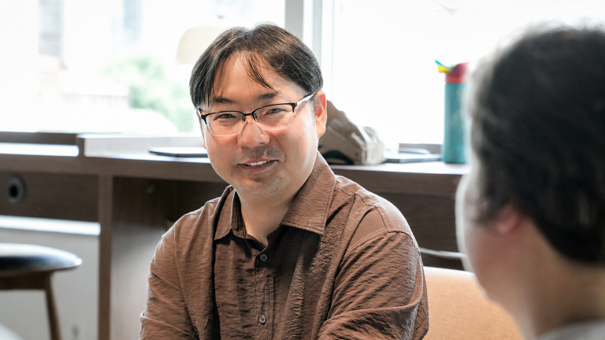 Man with glasses in a brown shirt talks to a person facing away at a table.