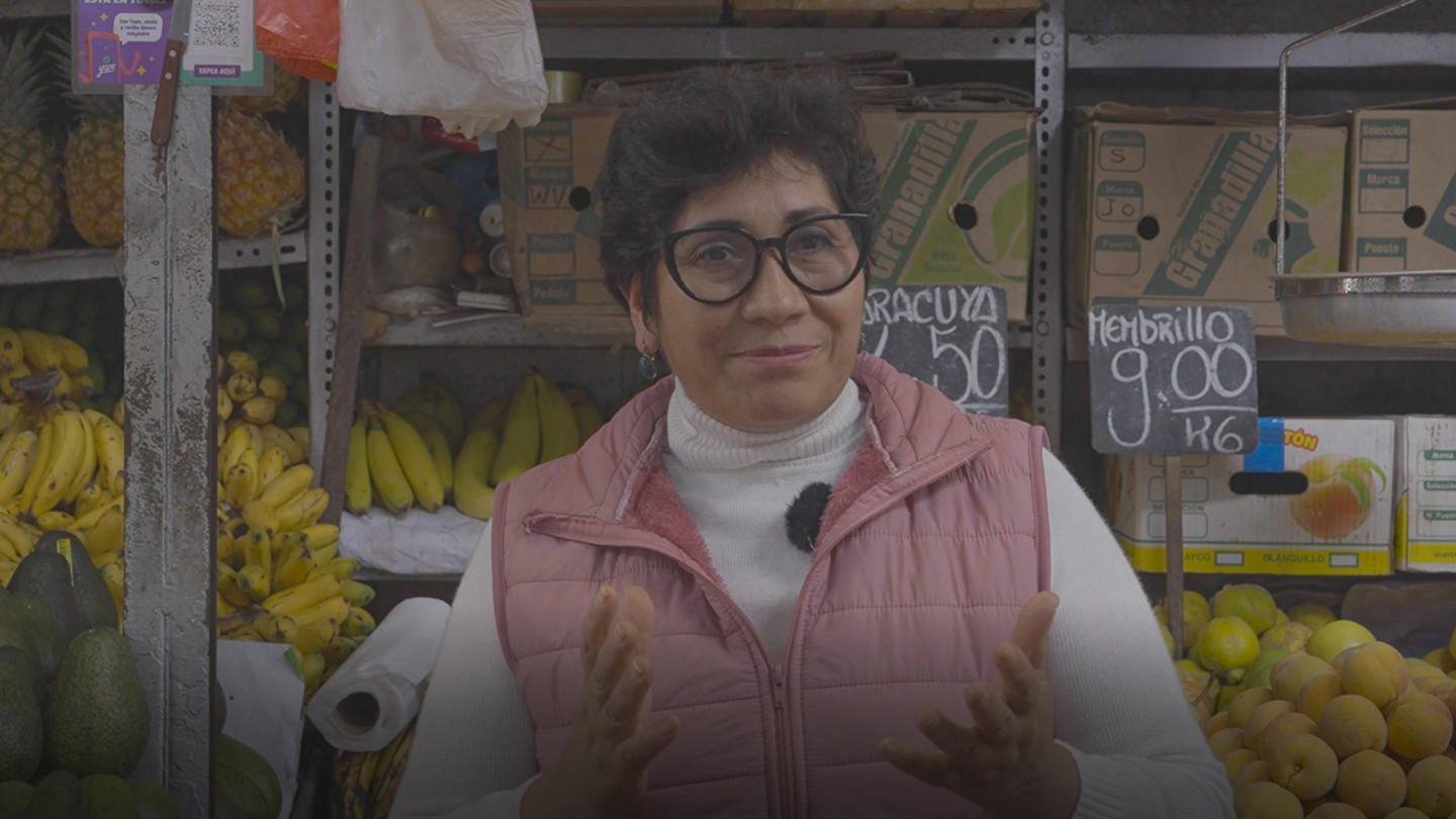 Market vendor wearing pink vest and glasses, gesturing amid fruit crates.