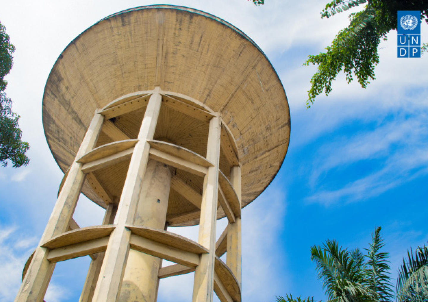 Low-angle view of a tall concrete water tower with a circular top, framed by palm trees against a blue sky.