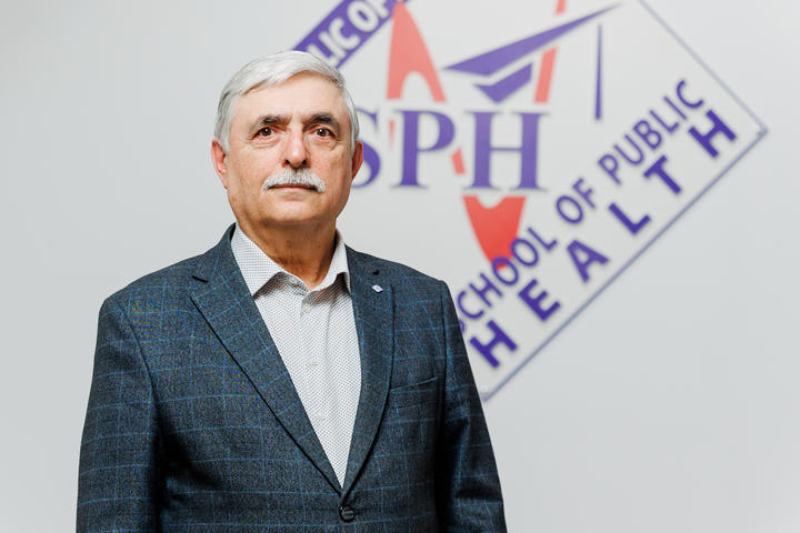 A serious-looking man with gray hair stands in front of a School of Public Health logo.