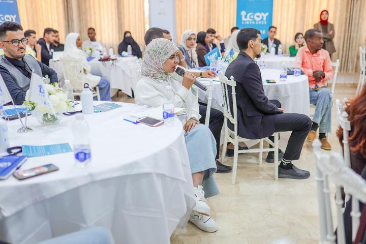 Attendees sit at round tables with water bottles in a bright hall during a formal event.