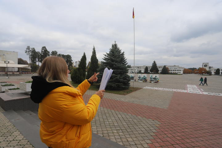Person in a bright yellow jacket reads from a paper in an outdoor plaza with a flagpole.