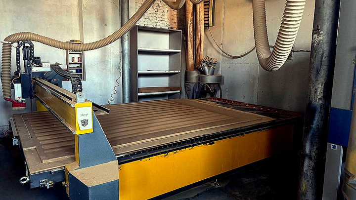 Industrial CNC cutting table in a workshop, yellow base, blue gantry, dust hose.