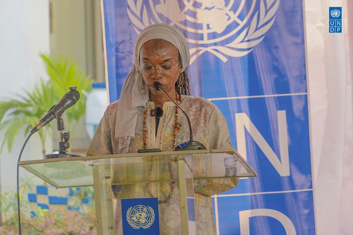 Photograph of a woman in hijab speaking at a UN podium with the UN emblem and blue backdrop.
