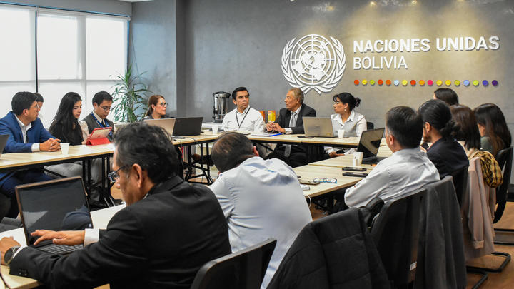 Delegates at a United Nations meeting in Bolivia with UN logo on the wall.