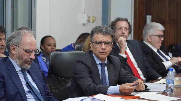 Group of suited men around a conference table; central figure speaks during a formal meeting.